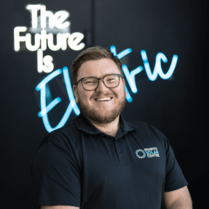 A man in a black shirt standing in front of a neon sign that says the future is electric.