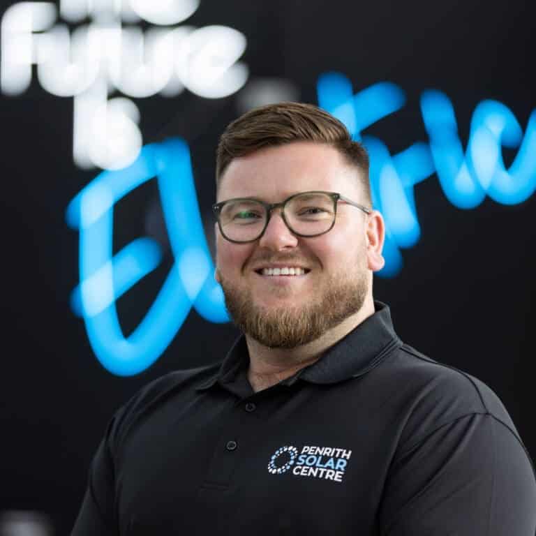 A photo of PSC owner and leader Jake Warner. He has a beard and glasses, and is smiling, standing in front of a neon sign that reads "The future is electric".