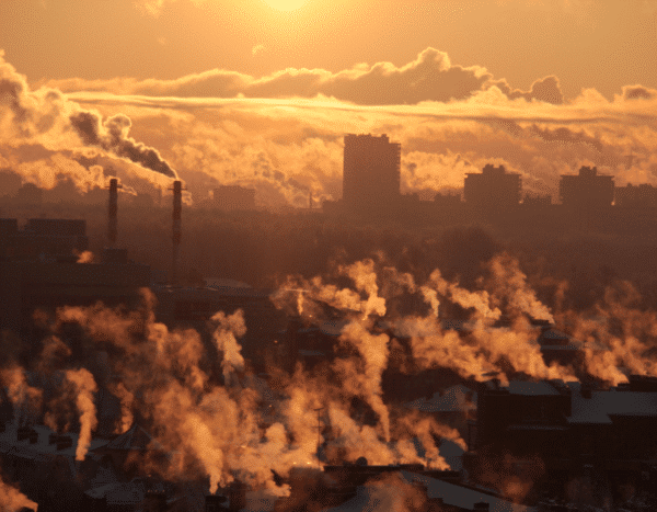Sunrise over a city skyline with silhouetted buildings and smokestacks emitting steam against a cloud-filled, orange sky.