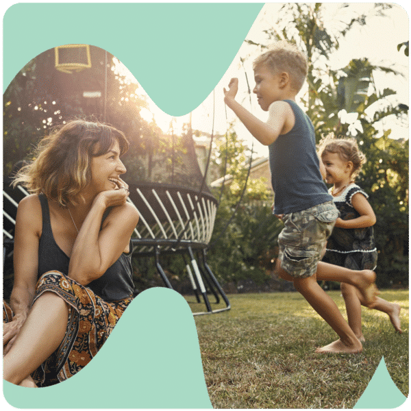 A woman smiles watching two children play with a toy airplane in a sunlit backyard.