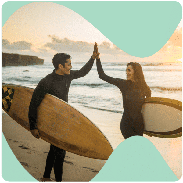 Two surfers in wetsuits high-fiving on the beach at sunset, both holding surfboards, with ocean waves in the background.
