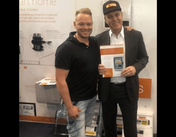 Two men posing with a document, one of them wearing a hat, standing in a booth with electronic products on display.